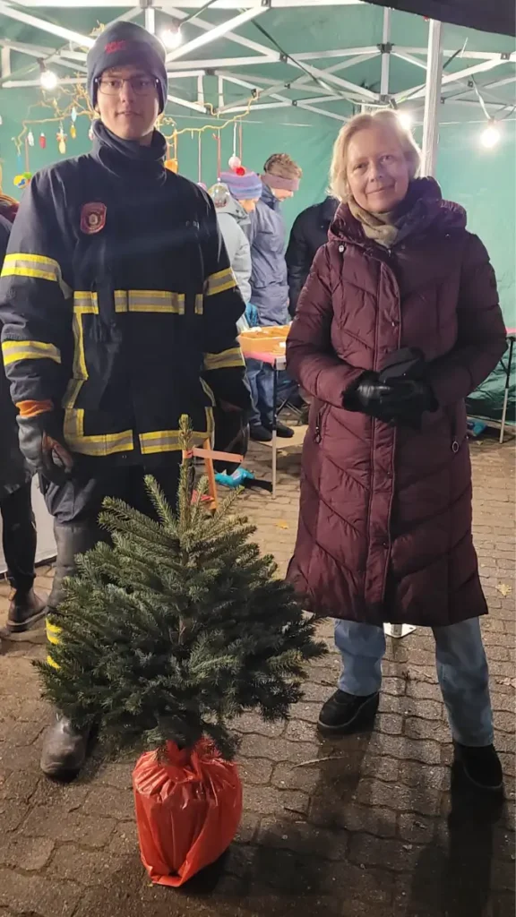 Zwei Menschen schauen in die Kamera. Die linke Person trägt eine Feuerwehruniform. Die andere Person ist winterlich gekleidet. Vor den beiden Personen steht ein kleiner Tannenbaum im Topf.