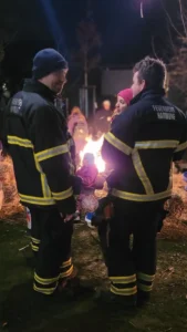 Zwei Personen in Feuerwehruniformen schauen zu einer Feuerschale.