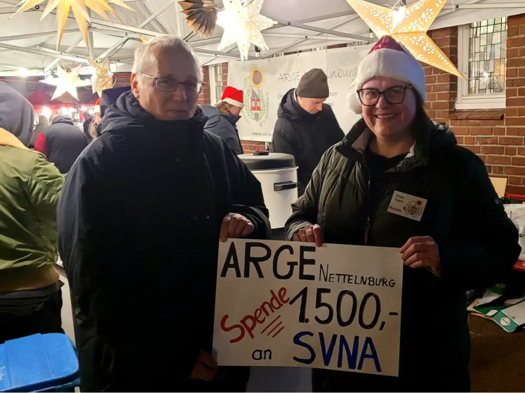 Zwei Personen schauen in die Kamera. Beide halten einen symbolischen Scheck mit der Aufschrift "ARGE Nettelnburg. Spende 1.500 Euro an SVNA. Im Hintergrund weitere Menschen, die an einem Glühweinstand tätig sind.