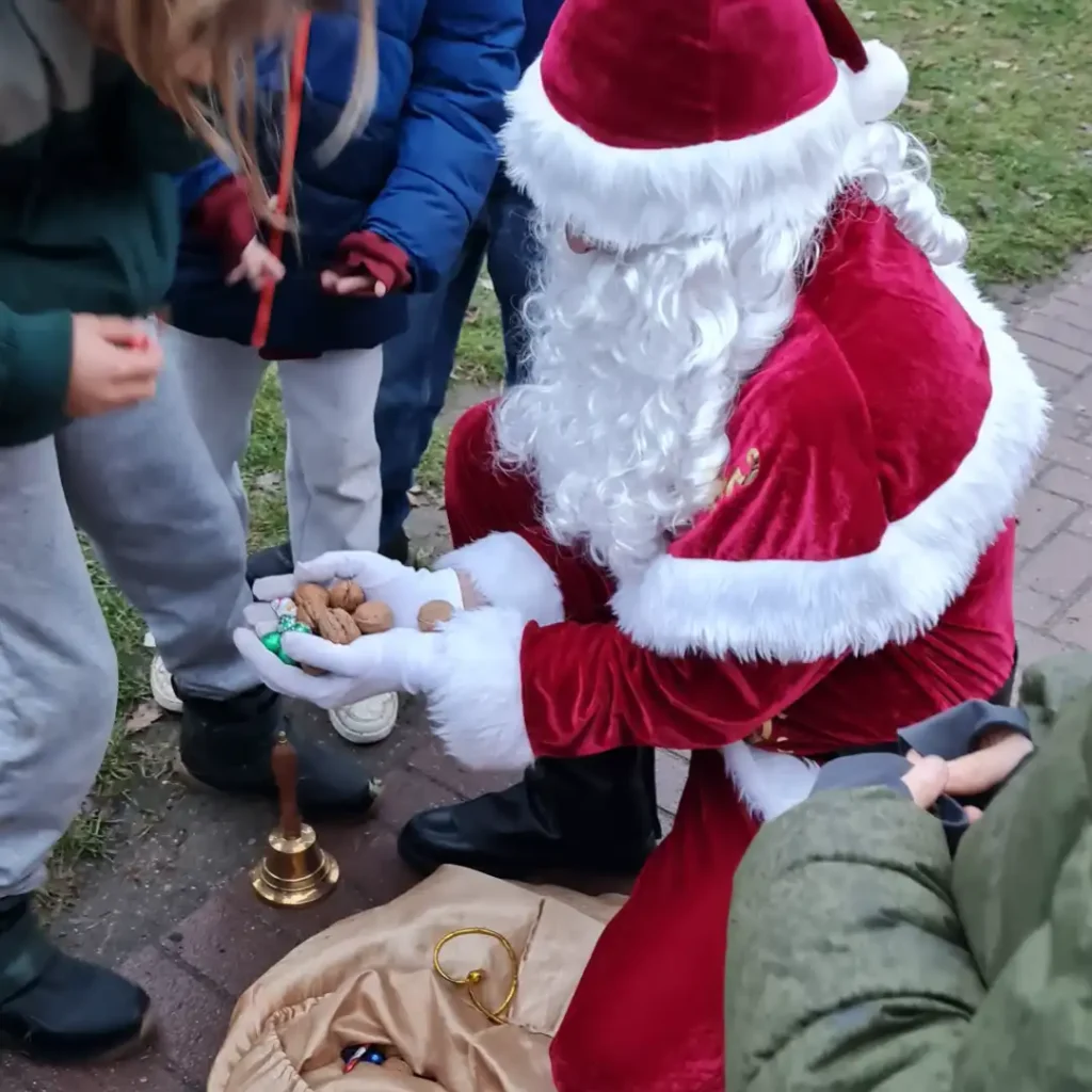 Das Bild zeigt einen hockenden Weihnachtsmann, der ein paar Nüsse in den Händen hält und von einigen Kindern umringt ist.